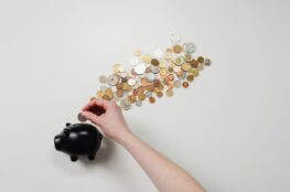 Hand inserting a silver coin into a black piggy bank beside scattered coins