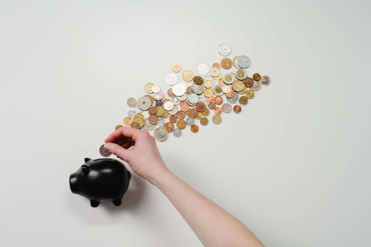 Hand inserting a silver coin into a black piggy bank beside scattered coins