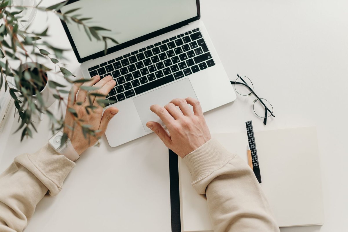 Overhead view of hands typing on a laptop beside reading glasses, a notebook and a plant