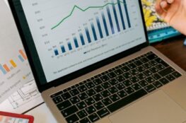 Laptop showing a quarterly revenue bar and line chart, with a hand holding a pen beside it and printed charts on the desk