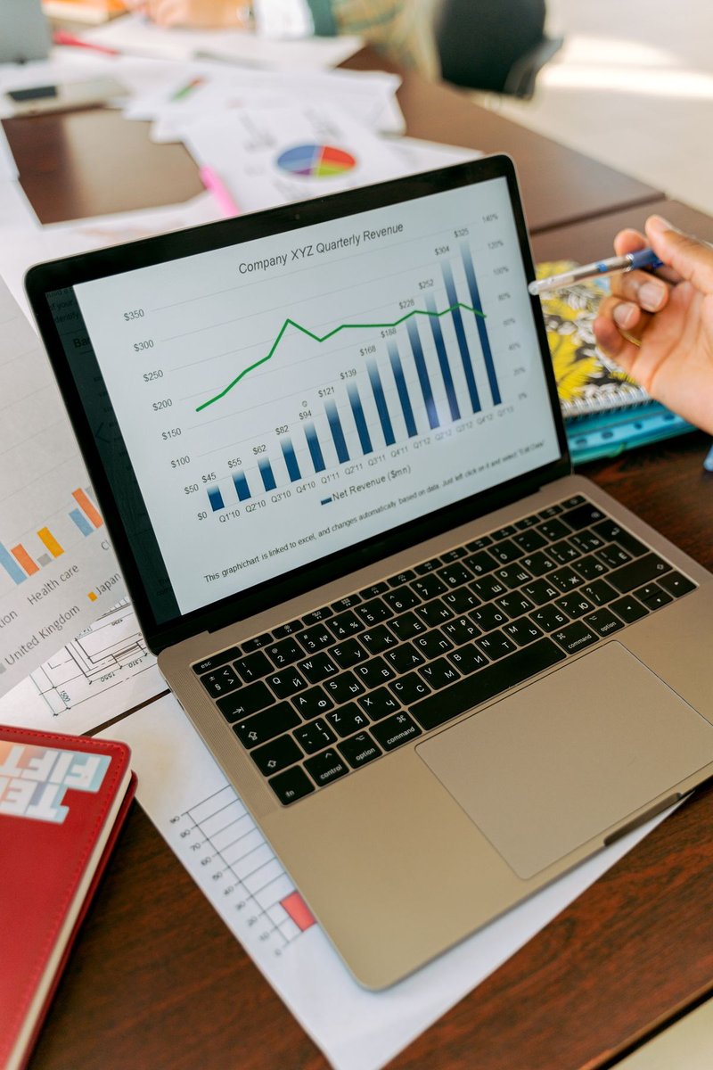 Laptop showing a quarterly revenue bar and line chart, with a hand holding a pen beside it and printed charts on the desk